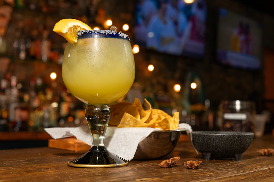 Salt-rimmed margarita with an orange wedge and a basket of chips on the bar